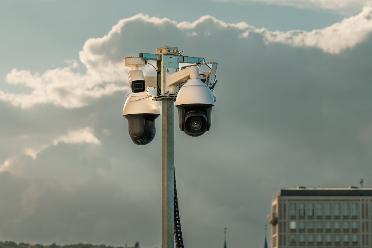 Services Two modern surveillance cameras mounted on a pole in an urban setting under a cloudy sky.
